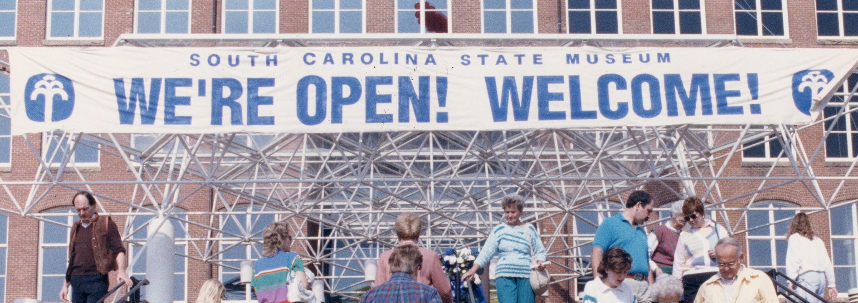 Group of people walking up stairs towards a large brick building with a banner over the entrance that reads "We're Open! Welcome!"