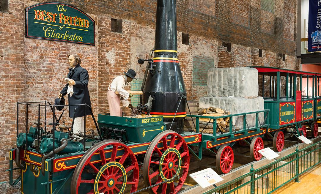 Full size train replica of The Best Friend of Charleston at the State Museum. Shows green train with red wheels and black smoke stack. 